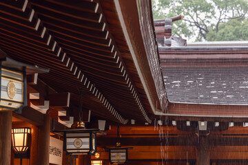Rainwater cascades from the wooden eaves of Meiji Shrine in Tokyo, highlighting traditional Japanese architecture. Ornate lanterns with chrysanthemum emblems softly illuminate the shrine interior