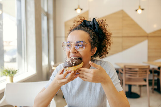 Portrait of a hungry young woman sitting in fast food restaurant and eating delicious doughnut.