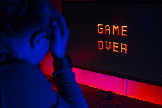 A guy sits at a computer in a dark room with neon lights on the monitor with the inscription game over