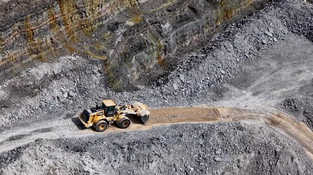 Aerial view of an open pit mining site shows a massive dump truck navigating a dirt roadway between steep rocky walls under bright daylight
