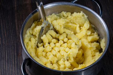 Fresh mashed potatoes mashed with a metal masher in a saucepan on a wooden table, closeup