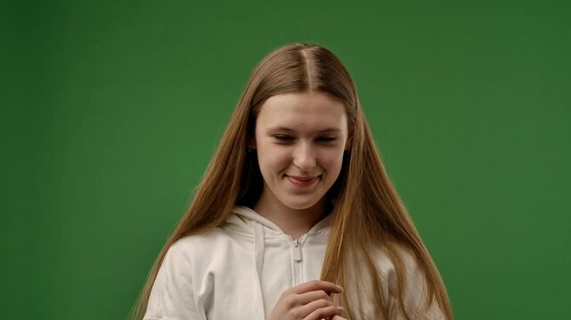 Teenage girl posing at camera smiling and touching hair, shy positive expression, isolated on chroma key green screen studio background mockup. Slow motion