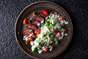 Sliced steak with Caesar Salad in black plate on black background.