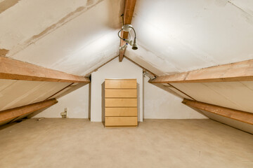 A bright attic space featuring wooden beams, a light beige carpet, and a simple dresser against the...