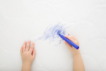 Baby hand holding marker and drawing blue scratches lines on white mattress cloth. Closeup. Draw of...