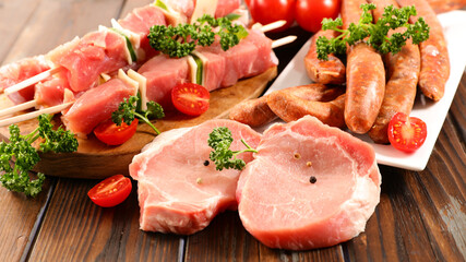 raw pork chop, sausage and skewer with cherry tomato and parsley on wooden table, ready for grilling, barbecue preparation scene with fresh meat