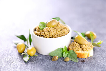 green olive tapenade in white bowl with toast, olive and basil on gray stone background, healthy mediterranean appetizer with soft natural light
