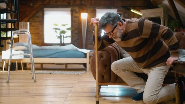Helpless lonely old 60s man falls to the floor while getting up from the couch leaning on a walking stick, elderly mobility problems pain, and stiffness in legs and back.