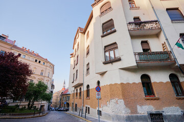 A street view of a European city with historic buildings and a church tower in the background. The architecture features ornate balconies and textured facades under a clear blue sky.