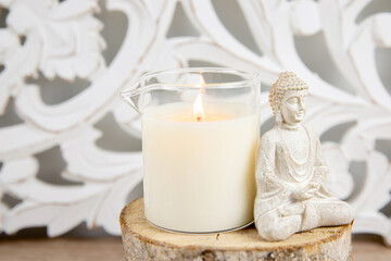Scented homemade massage oil candle in glass cup with pouring spout.  Bohemian style background, sitting Buddha figurine for decoration.