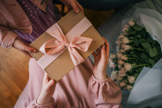 Close-up of a child giving a woman a gift box with a pink ribbon and a bouquet of delicate roses in soft natural light