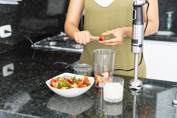 Woman preparing strawberry smoothie with hand blender in kitchen