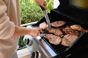 Woman’s hands checking grilled steaks on outdoor gas BBQ using knife and tongs. Juicy meat with sear marks. Summer cooking, backyard barbecue, lifestyle and food photography.
