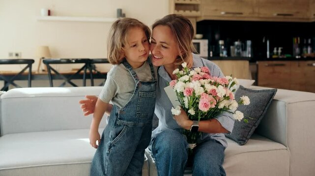 Young boy gives his mother a greeting card and flowers at home in a cozy setting. She reacts with joy and warmth. Concept of Mother's Day celebration and everyday family love. Mid-range shot natural