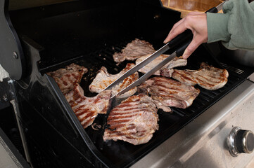 Hand holding tongs flipping grilled meat steaks on outdoor gas barbecue. Juicy, seared beef with grill marks. Summer cooking, backyard BBQ scene, high-protein lifestyle.