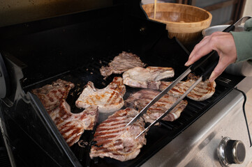 Hand holding tongs flipping grilled meat steaks on outdoor gas barbecue. Juicy, seared beef with grill marks. Summer cooking, backyard BBQ scene, high-protein lifestyle.
