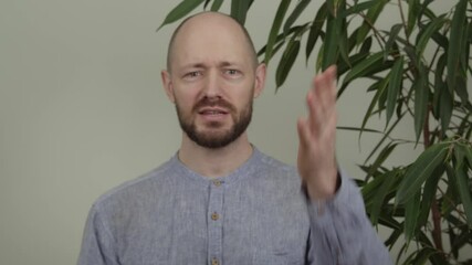 A guy blogger is giving a speech, looking at the camera, telling something funny, he is wearing a blue shirt and is in a studio next to a plant