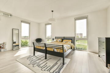 Spacious bedroom featuring large windows, natural light, minimalist decor, and a stylish bed with patterned bedding, creating a cozy atmosphere.