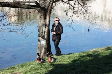 Young woman in sunglasses walking by the duck pond. Young girl walks by a lake in the autumn park. Adult female with short hair walks in on spring park by a river. Young woman resting near lake