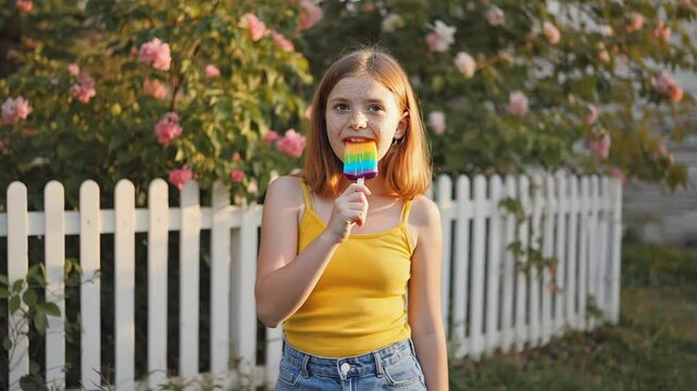 Girl in yellow top enjoying colorful rainbow popsicle in garden with blooming roses.

