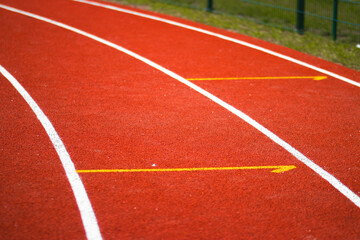 White lane markings on a red running track at an athletics stadium. The textured rubber surface and clear lines indicate starting positions. Shot in daylight, outdoor setting.

