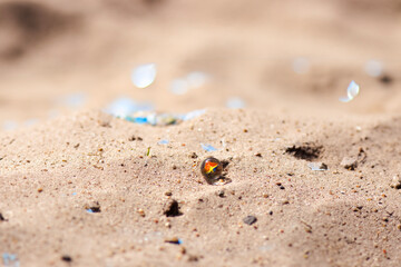 A glass ball with swirling multicolored paint lies on light beige sand. Vivid colors of red, blue, green, and yellow contrast with the soft, natural background of fine beach sand.

