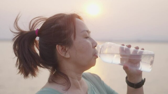 A close-up shows an overweight Asian woman in sportswear drinking water by the lakeside after exercising during sunset, a beginner caring for her health on her fitness journey.