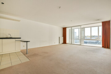 Spacious living room features large windows, beige carpet, and a minimalist kitchen setup, providing a bright and airy atmosphere.