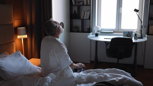 A middle-aged White man in his 50s, dressed in a t-shirt and pajama pants, sitting on the edge of his bed and waking up in the morning, turning on the lamp, as the sun shines in his window