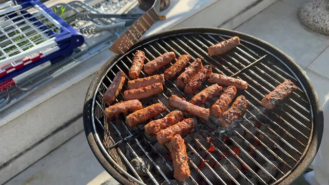 Ćevapi sizzling on hot charcoal grill, professional chef carefully turning meat sausages using metal spatula for even grilling during outdoor summer cookout