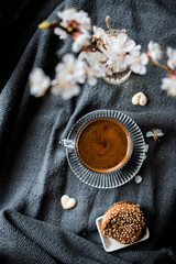 Coffee with a layer of foam is served in a clear glass cup on a matching saucer, placed on a textured gray blanket. White blossoms and small chocolates are scattered around. Top view, flatlay