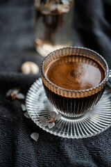 A glass cup of coffee sits on a ridged glass saucer atop a gray textured blanket. Nearby, a small vase holds delicate white flowers. A dish with sesame-coated cookies is visible
