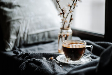 A glass cup of coffee sits on a ridged glass saucer atop a gray textured blanket. Nearby, a small vase holds delicate white flowers. A dish with sesame-coated cookies is visible