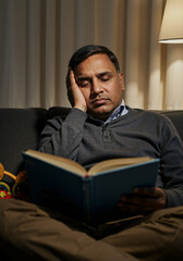 Tired man sitting on sofa falling asleep while reading a book at night