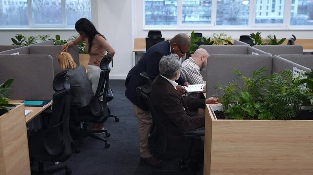 Diverse colleagues work in an office where some sit at desks with laptops and other devices while others stand. Large windows provide natural light, and the space includes plants and decor.