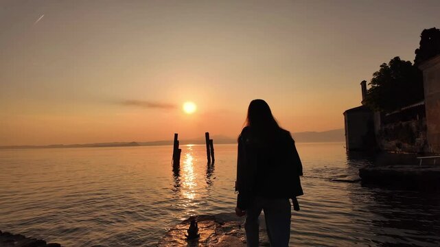 scenic video of a young woman in leather jacket running at the stone pathway on the bright golden hour sunset at Italian Garda lake 