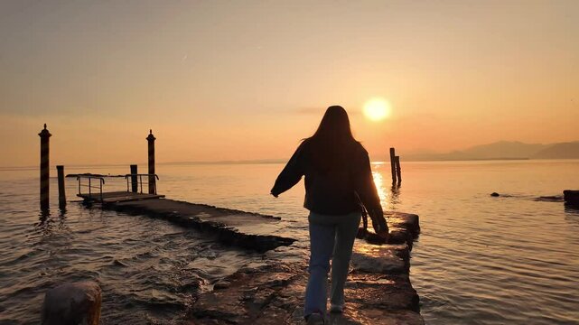 scenic video of a young woman in leather jacket running at the stone pathway on the bright golden hour sunset at Italian Garda lake 