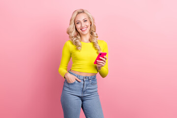 Young stylish woman in yellow top and jeans smiling while holding smartphone against a pink background