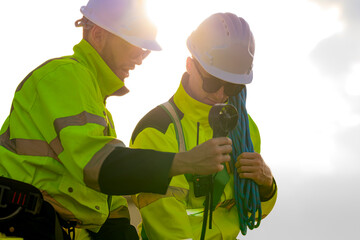 Two engineers wearing high visibility safety jackets and helmets measure wind speed with an anemometer and analyze data on a tablet at a renewable energy site. The cloudy sky adds depth to the scene.