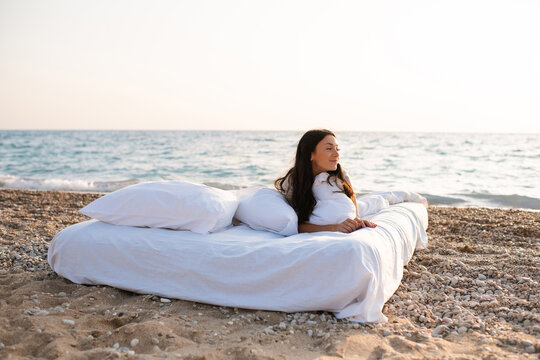 Woman lying in bed on white bedsheets over sea background. Summer vacation season.
