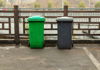 Naklejka premium Rubbish bin garbage square shape green and black with flip cover located next to fence in garbage disposal area. Old dirty trash cans plastic. Waste collection equipment in park.