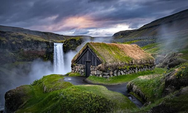 A dramatic close-up photograph of a solitary Icelandic turf house perched near the edge of a roaring waterfall