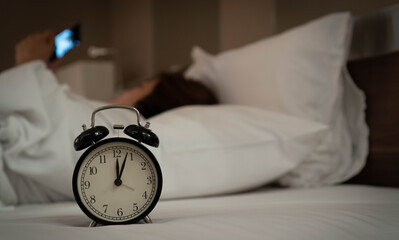 Vintage alarm clock showing midnight on a cozy bed, with a blurred person using a smartphone in the background. Late night habits, insomnia, and screen time before sleep concept