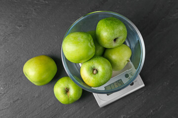 Electronic kitchen scale with bowl of green apples on dark textured table, flat lay