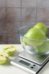 Electronic kitchen scale with bowl of green apples on white wooden table against tiled wall, closeup