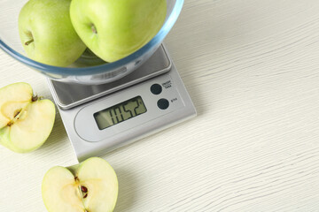 Electronic kitchen scale with bowl of green apples on white wooden table. Space for text