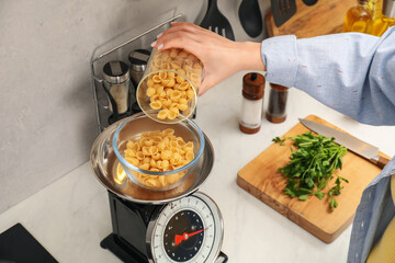 Woman adding raw pasta into bowl on mechanical kitchen scale at white marble countertop indoors,...