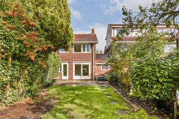 This serene garden setting features a two-story house with lush greenery, well-maintained lawn, and vibrant flora, creating a peaceful residential atmosphere.