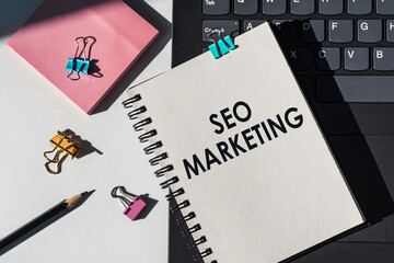 Close-up of white notebook with SEO MARKETING in black, on white background. Black laptop keyboard,...