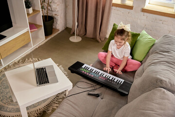 Little girl practicing keyboard on couch while watching online music class on laptop. Child...